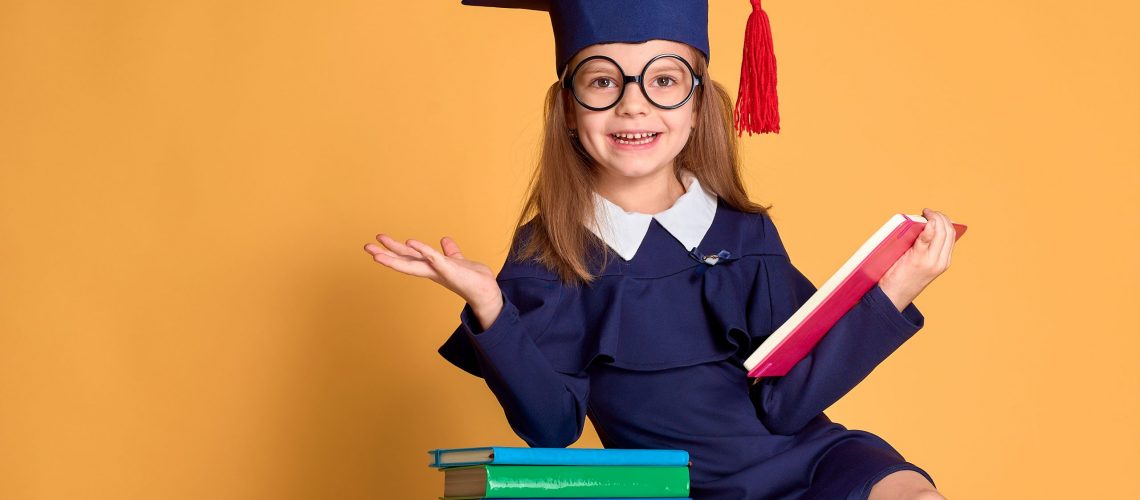 Excited smiling schoolgirl in glasses and graduation clothes cheering while sitting at desk with colourful books and pen over yellow background