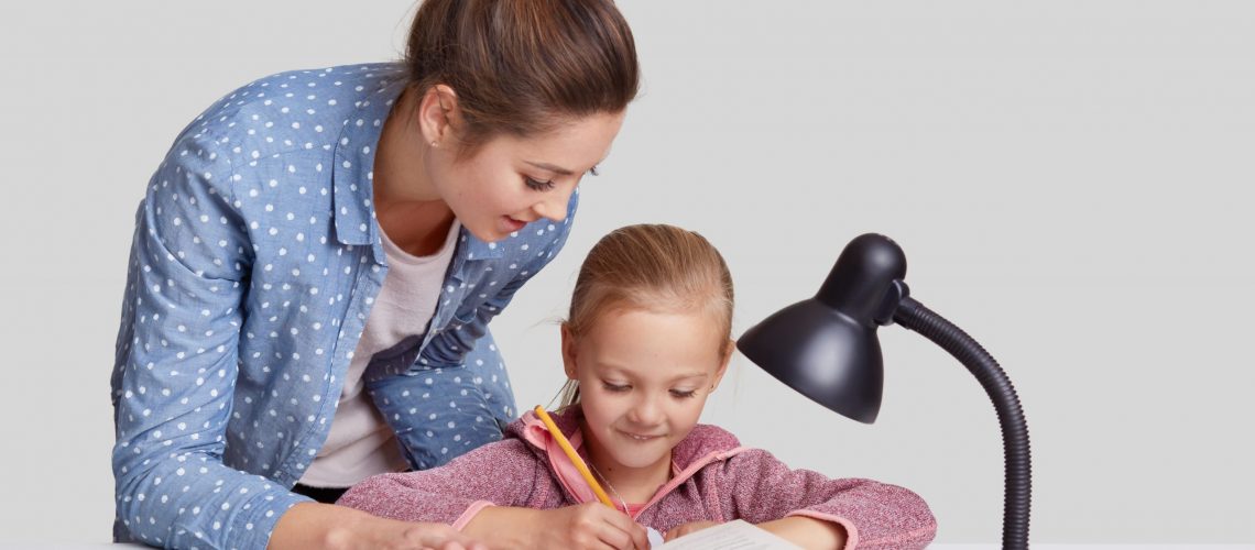 Small child rewrites information in notebook, has pleased expression, her mother stands near, tries to encourgae daughter for studying, help and explains material, isolated over white background