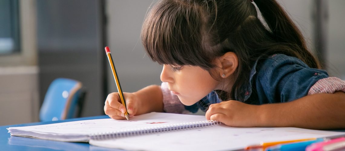 Focused black haired Latin girl sitting at school desk and drawing in her copybook. Education or back to school concept